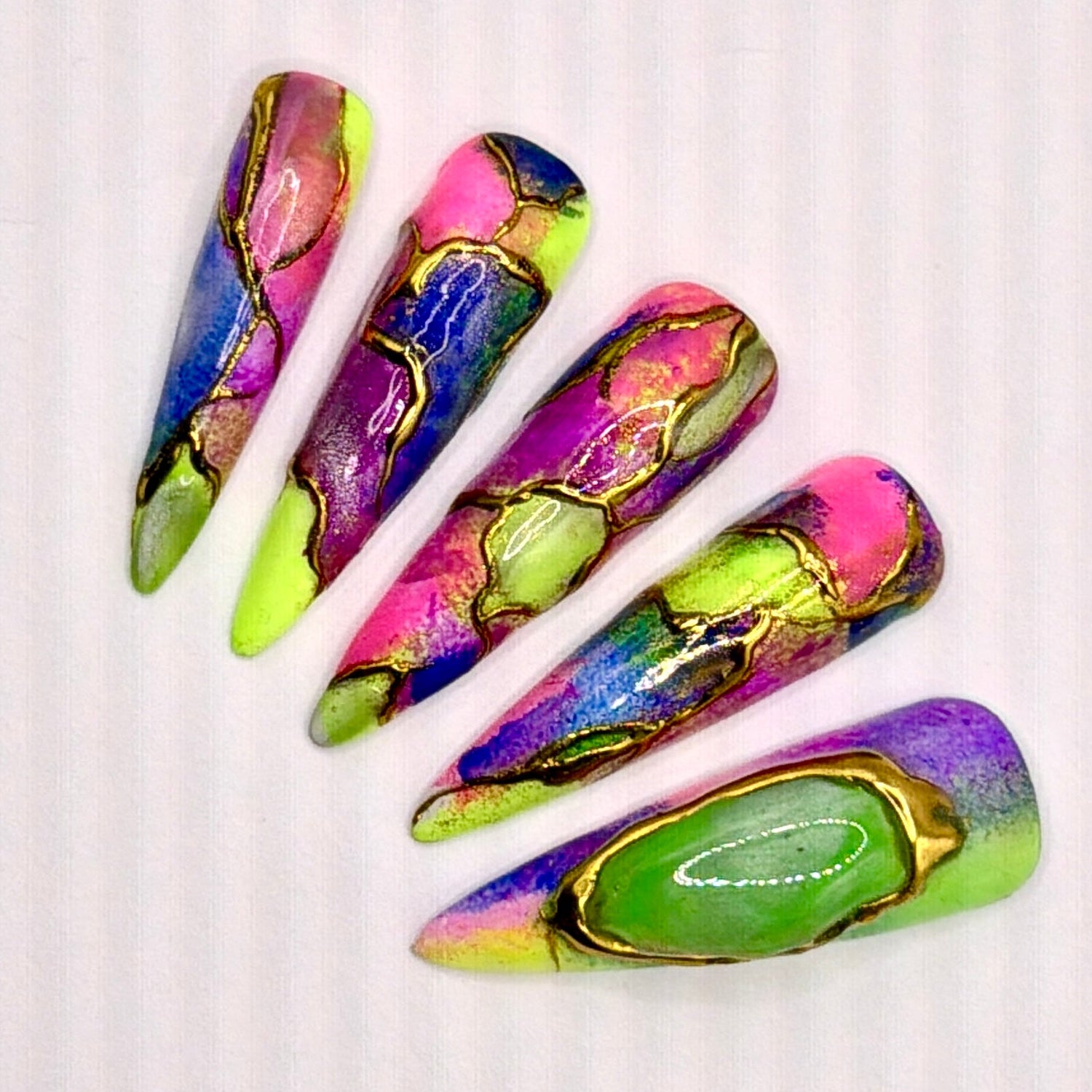 Five long stiletto-shaped press-on nails with a vibrant neon marbled pattern in pink, green, purple, blue, and yellow, arranged on a plain off-white background. glow in the dark