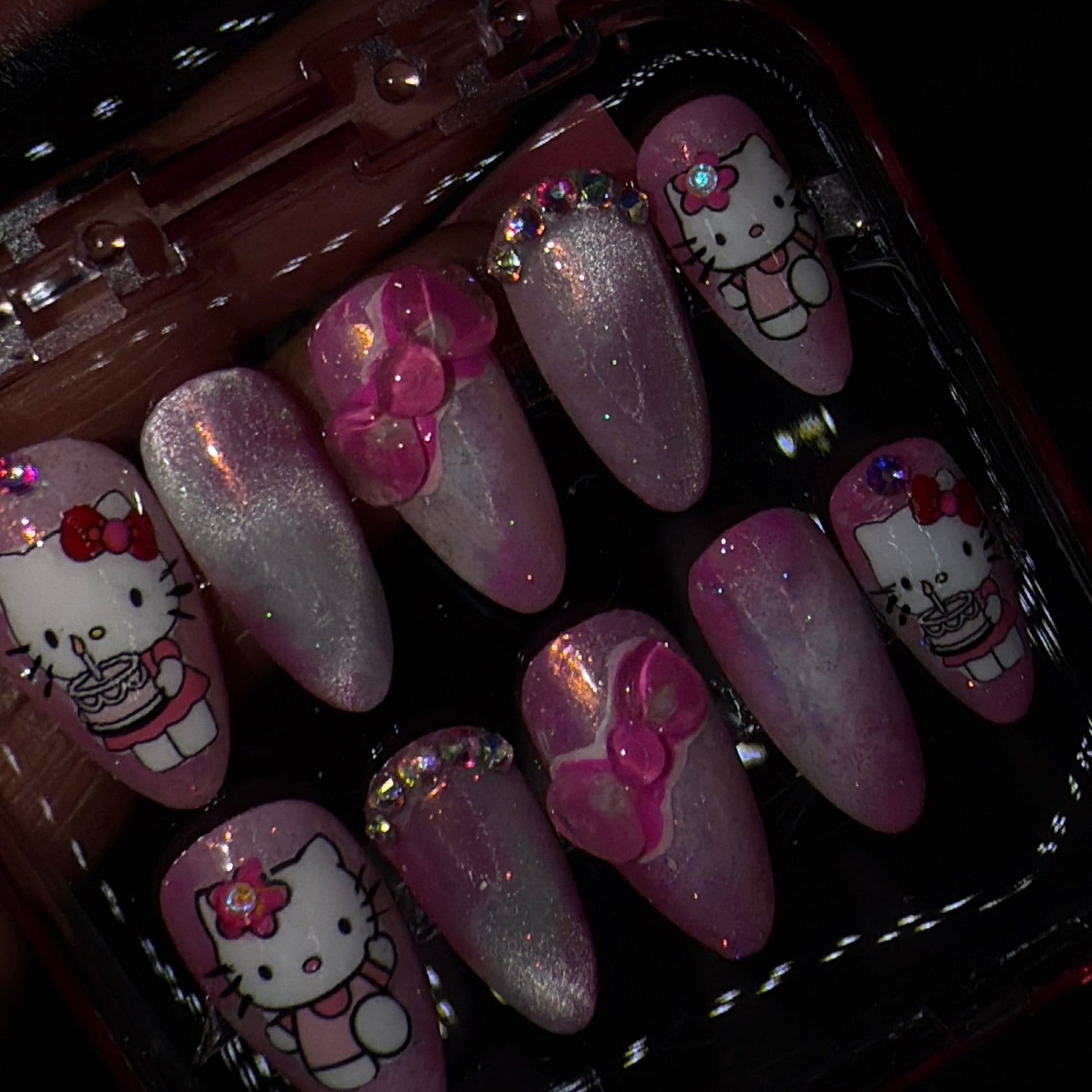 A set of pink and white Hello Kitty press-on nails displayed in a clear plastic case against a dark background. The nails feature different designs including Hello Kitty character prints, pink bows, and glittery pink tips.